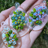 A hand holding three pendants made of colorful glass beads, each featuring a different flower design.