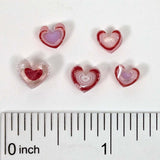 a collection of heart-shaped objects, possibly made of glass or crystal, arranged on a white surface. A ruler is placed below the objects to provide a scale reference.