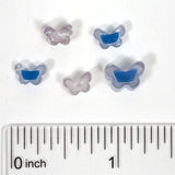 five small, clear beads in the shape of butterflies, with a ruler for scale placed below them.