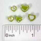 a collection of green heart-shaped objects, possibly beads or charms, placed next to a ruler for size comparison.