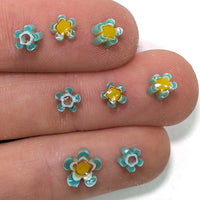 A close-up image of a person's fingers holding small, colorful flower-shaped objects made of a material that appears to be glass or a similar material.