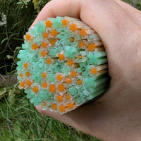 A hand is holding a circular arrangement of green and orange flower-shaped objects, which appear to be made of a material that resembles glass or plastic.
